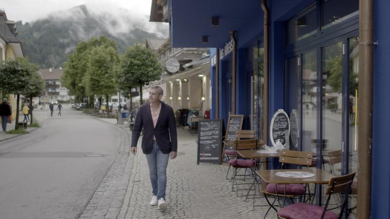 "Kaminer Inside: Kultursommer mit Hindernissen": Wladimir Kaminer läuft durch eine Straße in Oberammergau vorbei an einem Café mit Außenbestuhlung ohne Kunden.