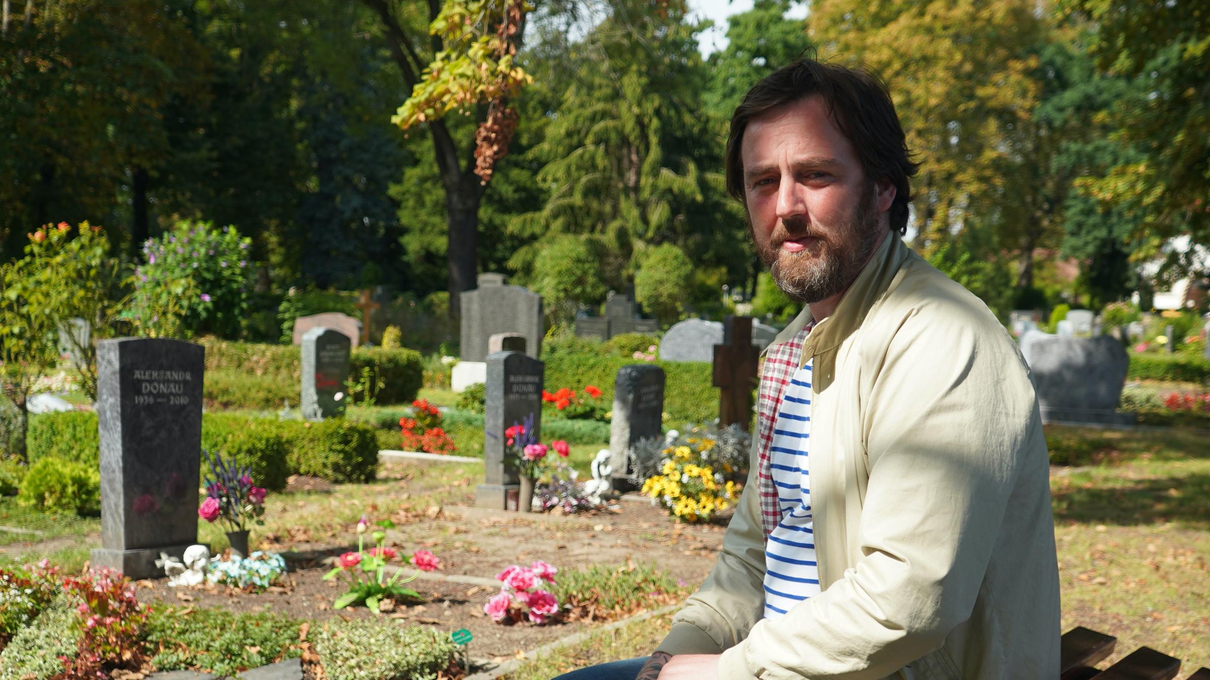 "37°: Mehr als Sarg und Service - Die neuen Bestatter*innen" Eric sitzt auf einem Friedhof vor Gräbern.