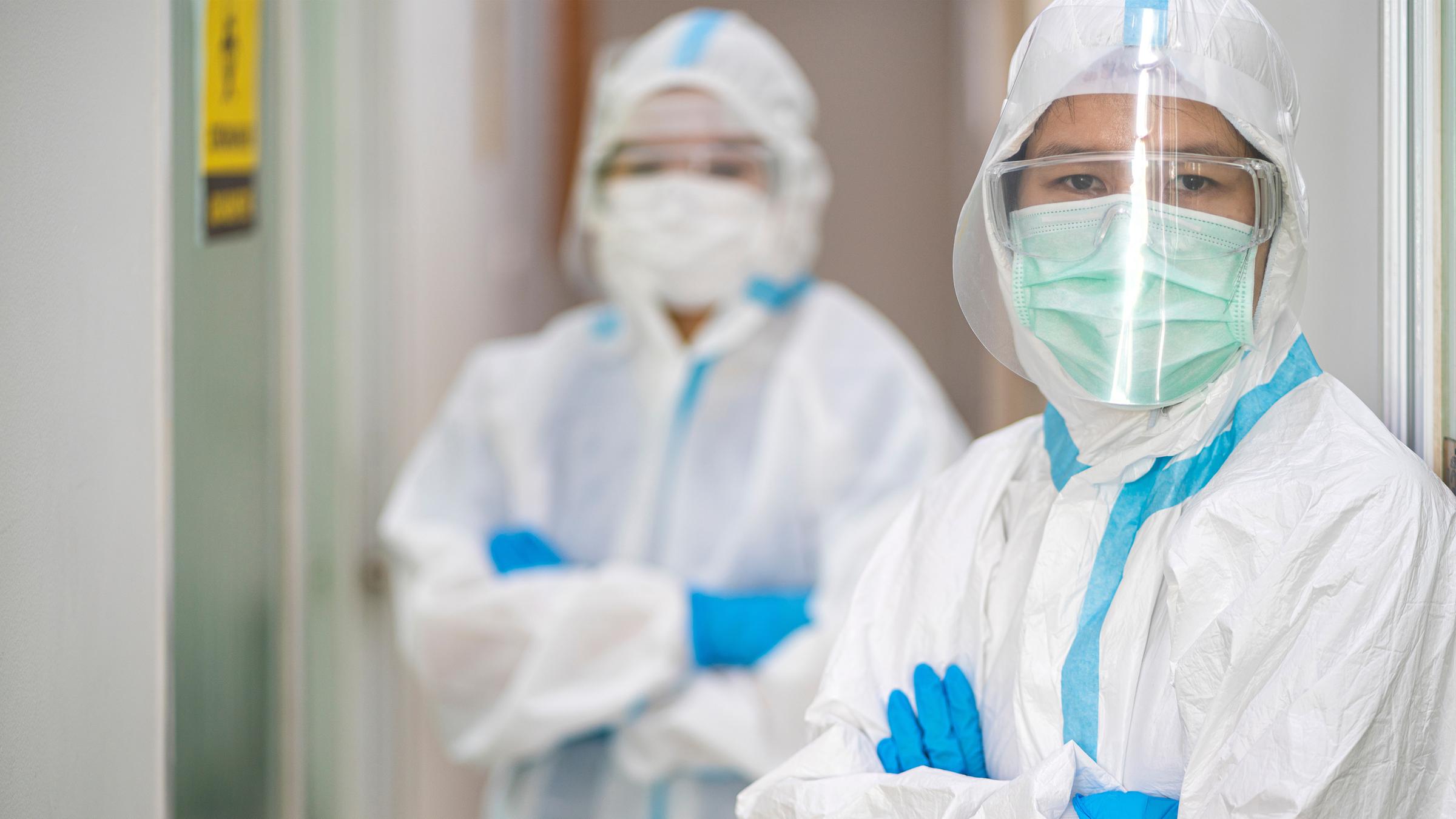 "Corona 2020 - Schock und Versagen": Zwei Personen tragen weiße Schutzkleidung und schauen mit verschränkten Armen in die Kamera.