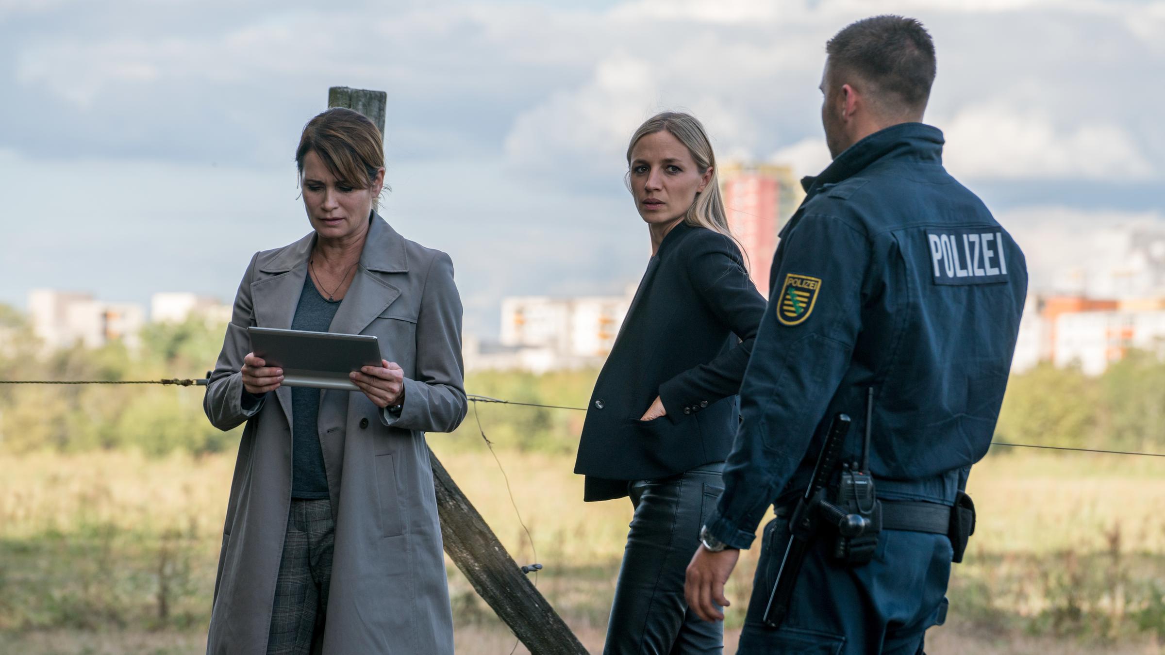 "Das Quartett – Das Mörderhaus": Maike (Anja Kling), Pia (Annika Blendl) und ein Polizist stehen vor einer Wiese. Der Polizist ist nur von hinten zu sehen. Pia wirft ihm einen Blick über die Schulter zu. Maike sieht auf ihr Tablet.
