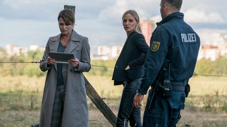 "Das Quartett – Das Mörderhaus": Maike (Anja Kling), Pia (Annika Blendl) und ein Polizist stehen vor einer Wiese. Der Polizist ist nur von hinten zu sehen. Pia wirft ihm einen Blick über die Schulter zu. Maike sieht auf ihr Tablet.