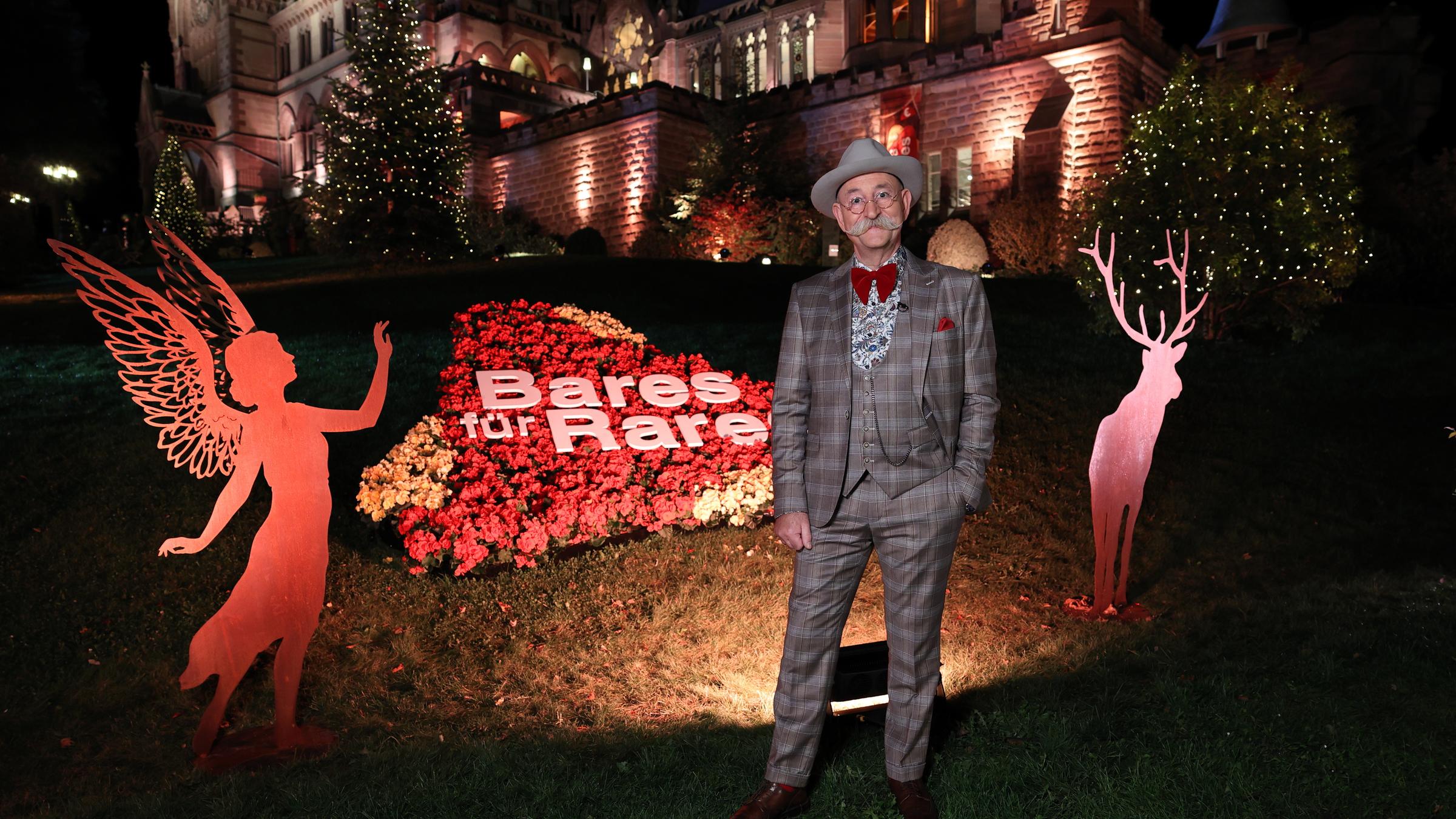 "Bares für Rares - Deutschlands größte Trödelshow":  Horst Lichter steht  nachts vor dem Sendungslogo, das aus Blumen nachgebildet wurde. Im Hintergrund ist der weihnachtlich dekorierte Park von Schloss Drachenburg zu sehen.