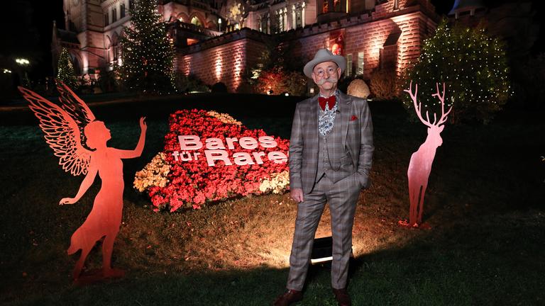 "Bares für Rares - Deutschlands größte Trödelshow":  Horst Lichter steht  nachts vor dem Sendungslogo, das aus Blumen nachgebildet wurde. Im Hintergrund ist der weihnachtlich dekorierte Park von Schloss Drachenburg zu sehen.