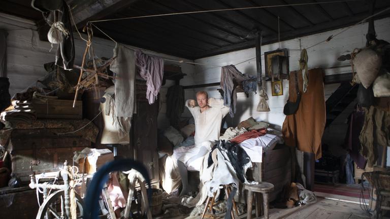 "Die Teufelsfeder": Wir sehen den Schneider (Jan Budař) – noch im weißen Schlafanzug – auf seinem heimischen Bett. Das Zimmer, in dem das Bett steht, sieht ärmlich und unordentlich aus. Überall verstreut liegen Kissen und Kleider, Tücher, Decken, ein Spinnrad, ein paar heruntergekommene Möbelstücke. Die aufgehende Sonne erleuchtet das Zimmer, macht es aber dadurch nicht wohnlicher.