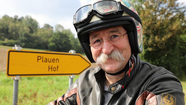 Horst Lichter sitzt auf einem Motorrad. Er hat einen Motorradhelm auf. Im Hintergrund steht ein Straßenschild mit der Aufschrift Plauen und Hof.