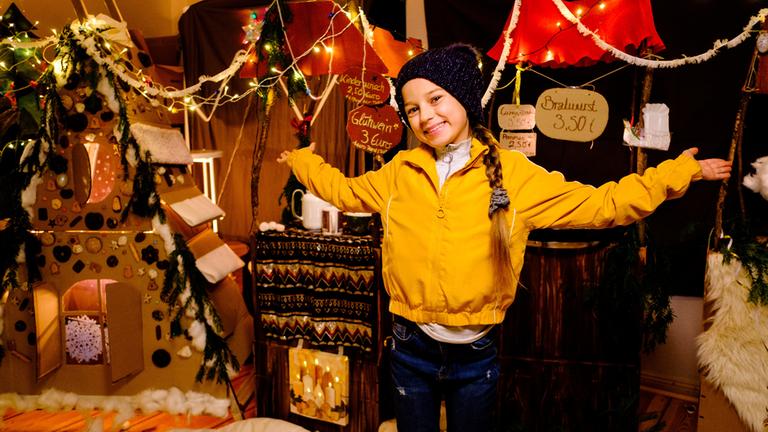 "Liebe. Jetzt! Christmas Edition - Weihnachtszauber": Das Mädchen Martha (Úna Lir) steht in einer gelben Jacke in einem selbst gebastelten Weihnachtsmarkt und breitet die Arme aus.