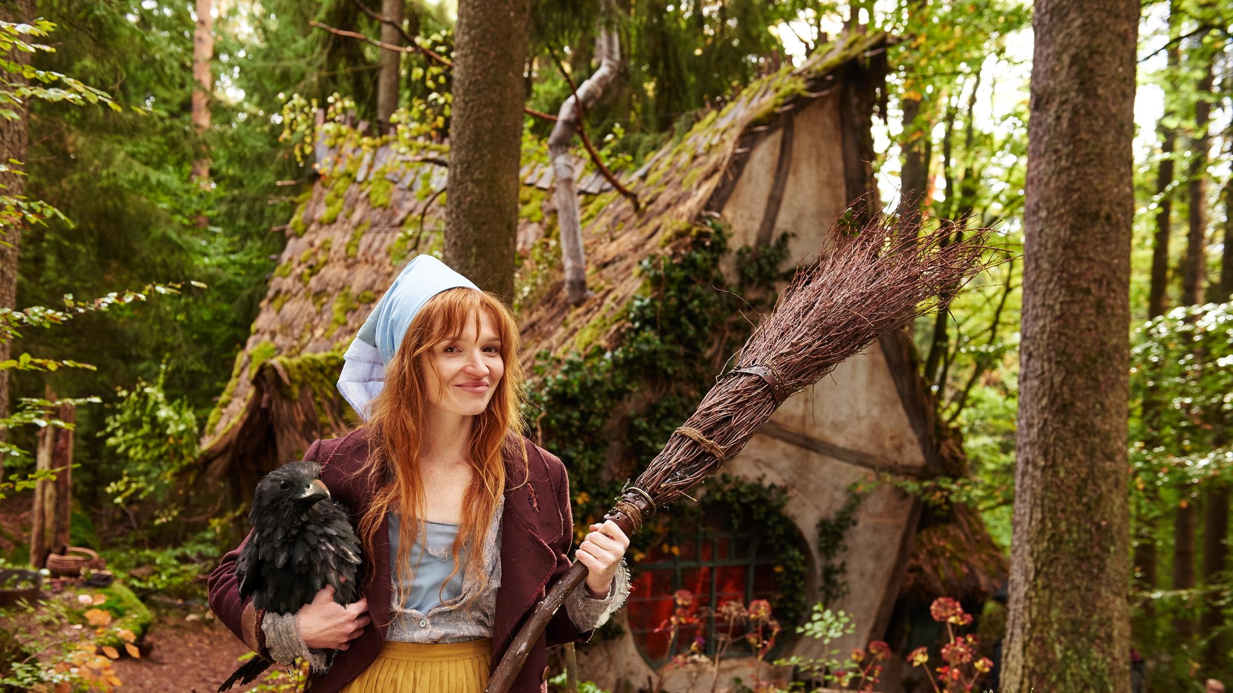 "Die kleine Hexe": Die kleine Hexe (Karoline Herfurth) spaziert mit dem Besen in der Hand und dem Raben unterm Arm durch den Wald. Hinter ihr ist das kleine, krumme und schiefe Hexenhaus zu sehen.