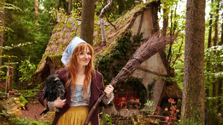 "Die kleine Hexe": Die kleine Hexe (Karoline Herfurth) spaziert mit dem Besen in der Hand und dem Raben unterm Arm durch den Wald. Hinter ihr ist das kleine, krumme und schiefe Hexenhaus zu sehen.