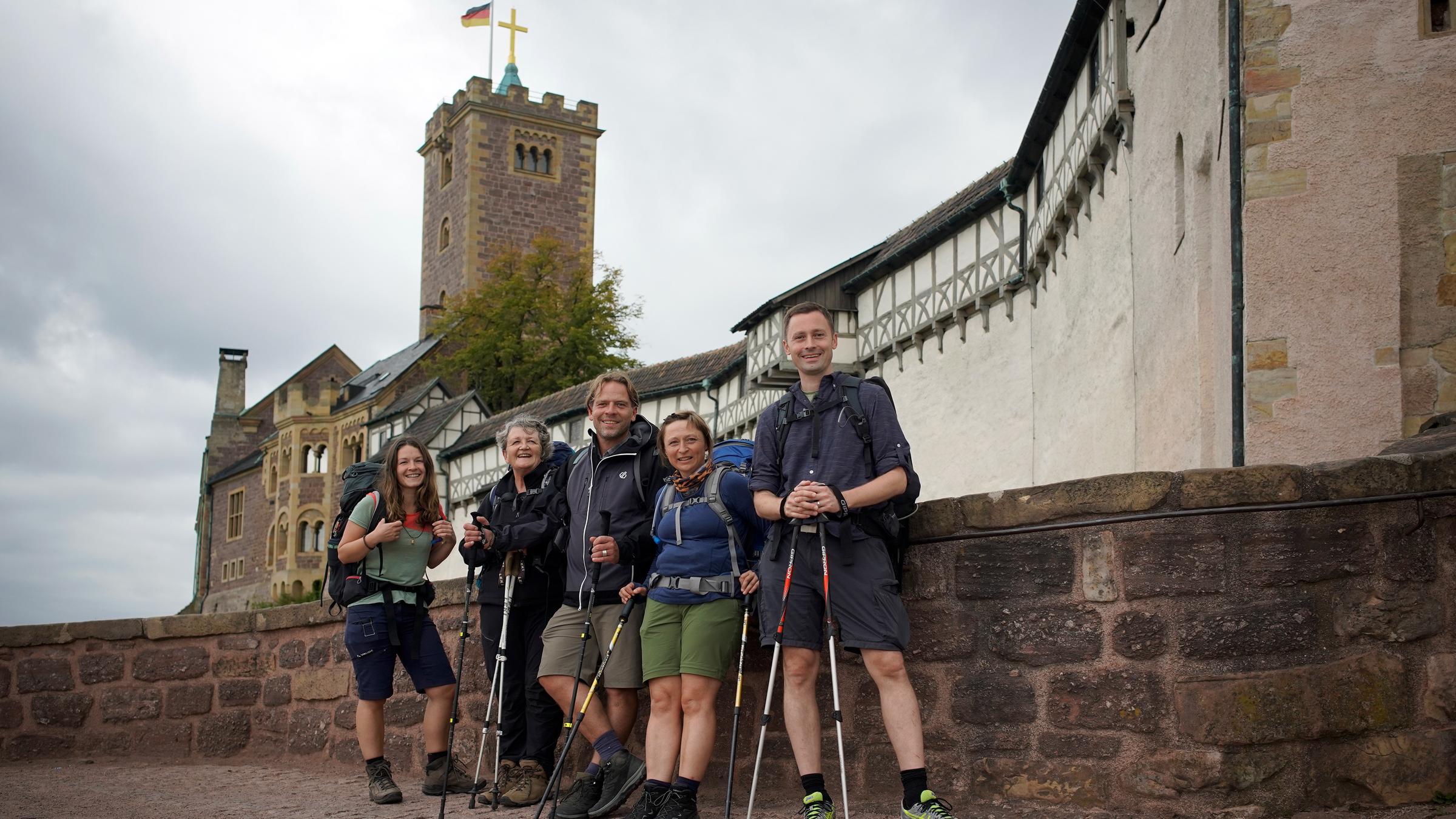 "Schritt für Schritt - Pilgern auf dem Lutherweg": Drei Frauen und zwei Männer stehen mit Outdoorkleidung, Walking-Stöcken und Rucksäcken vor einem Gemäuer, im Hintergrund eine Burgfestung. Sie schauen alle lächelnd in die Kamera