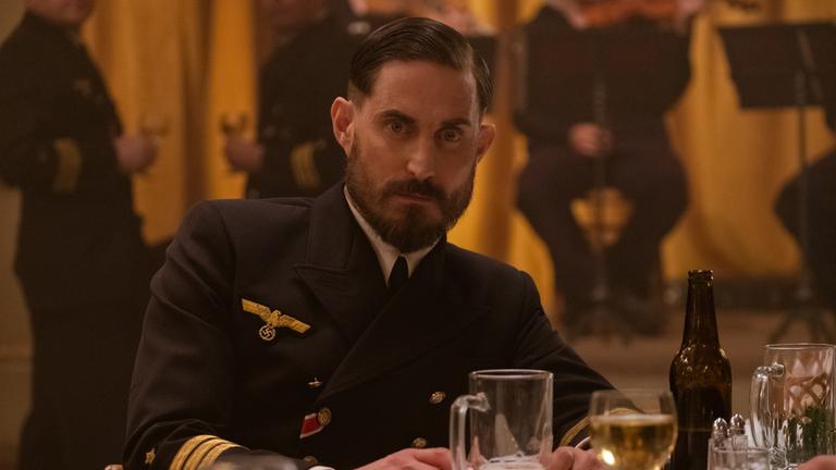 "Das Boot": Johannes von Reinhartz (Clemens Schick) sitzt an einem gedeckten Tisch und sieht nach vorn. Im Hintergrund sind verschwommen zwei Personen mit Weingläsern und eine Bühne mit Musikern zu sehen.