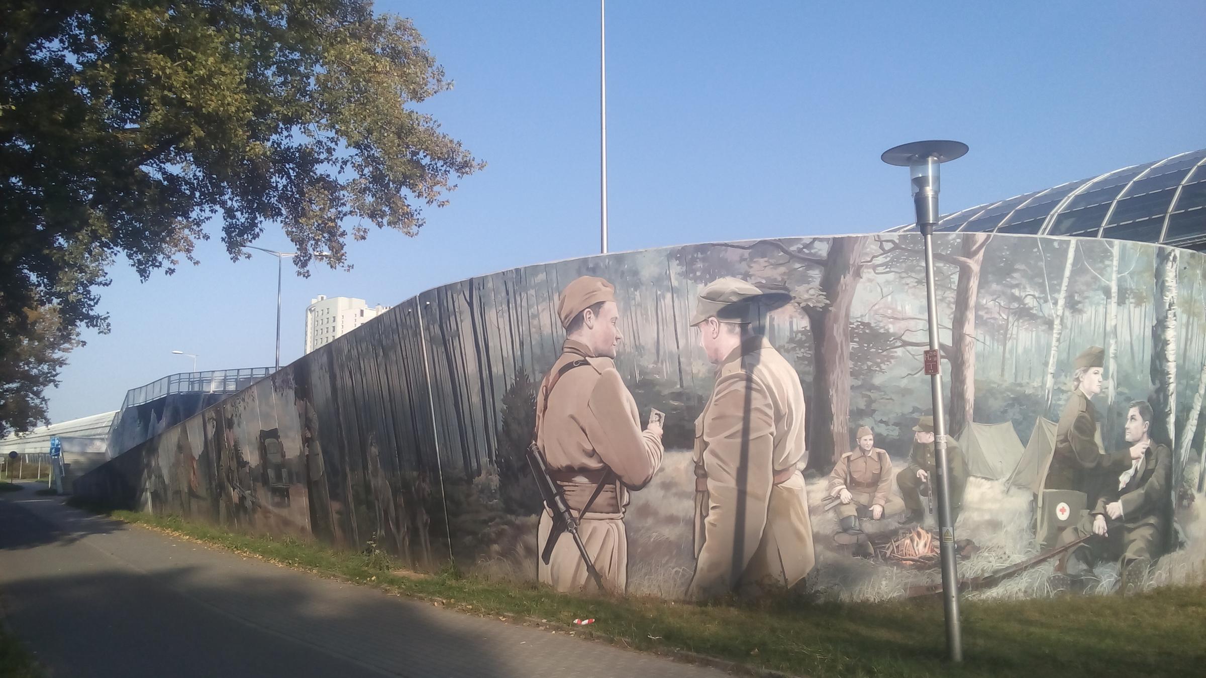 "Der Kniefall von Warschau – Die Macht der Erinnerung": Ein Gemälde auf einer großflächigen Mauer zeigt zwei junge polnische Soldaten in Uniform, sie betrachten das Foto eines jungen Mädchens.