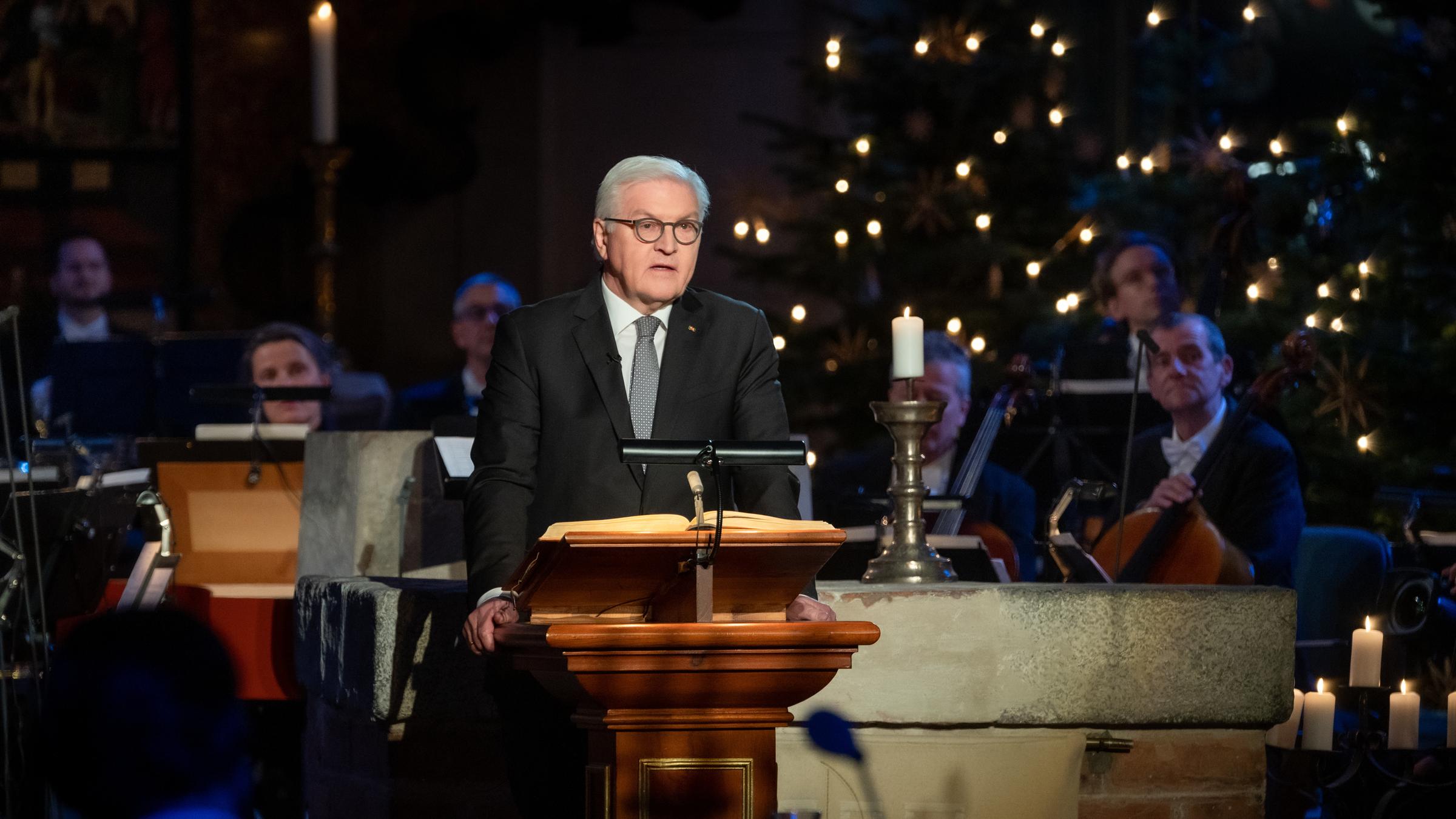 "Weihnachten mit dem Bundespräsidenten": Bundespräsident Frank-Walter Steinmeier steht an einem Pult und liest aus der Weihnachtsgeschichte. Im Hintergrund das Orchester und die Lichter eines Weihnachtsbaumes.