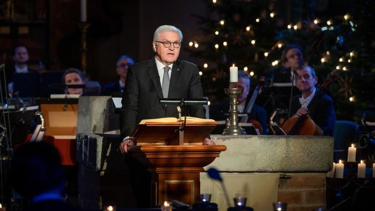 "Weihnachten mit dem Bundespräsidenten": Bundespräsident Frank-Walter Steinmeier steht an einem Pult und liest aus der Weihnachtsgeschichte. Im Hintergrund das Orchester und die Lichter eines Weihnachtsbaumes.