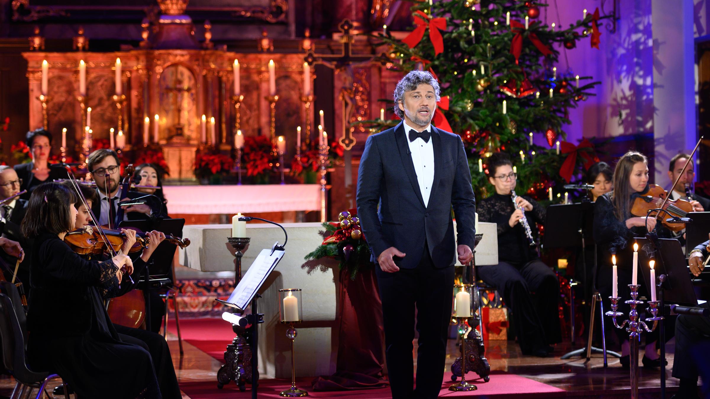 "Weihnachten mit Jonas Kaufmann": Jonas Kaufmann steht singend vor dem Orchester. Im Hintergrund der mit Kerzen dekorierte Altar und ein großer Weihnachtsbaum.