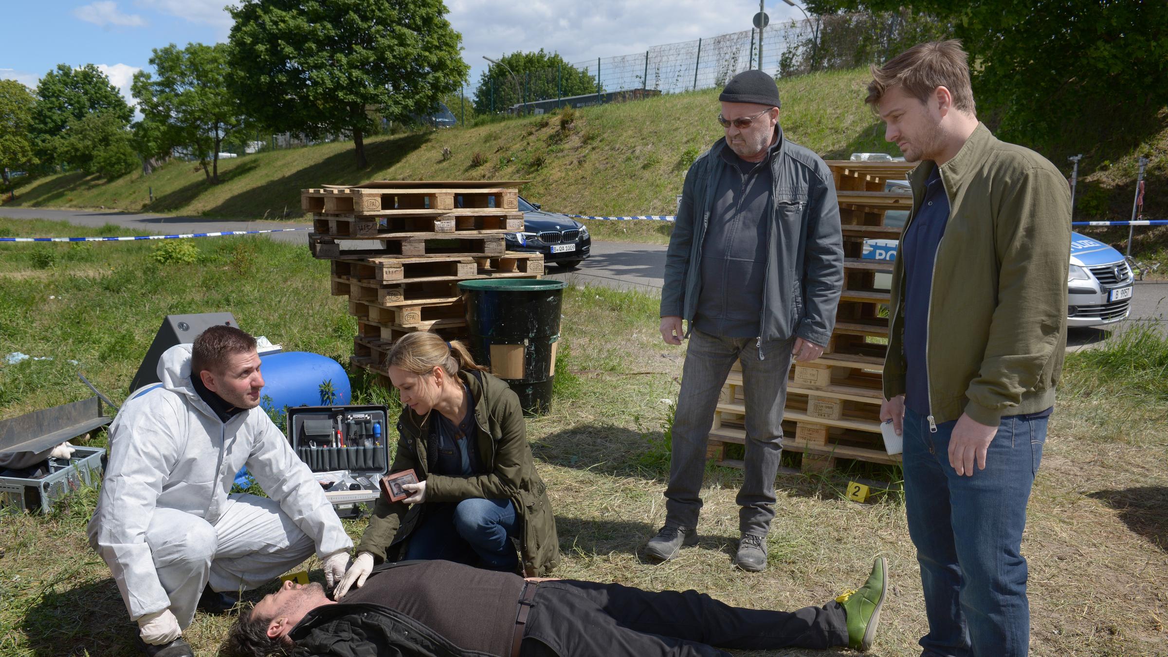 Ein starkes Team - Den Parkplatz bitte sauber halten": Linett (Stefanie Stappenbeck) kniet neben dem Opfer Prof. Thalhammer (Matthias Deutelmoser). Daneben stehen Otto (Florian Martens) und Sebastian (Matthi Faust) zwischen Paletten am Straßenrand.