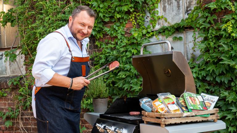 "ZDFzeit: Die Tricks der Lebensmittelindustrie - Turbo-Chips, Veggie-Burger & Co.": Produktentwickler Sebastian Lege steht am Grill, auf dem vegetarische Burger unterschiedlicher Hersteller liegen, und lacht in die Kamera.