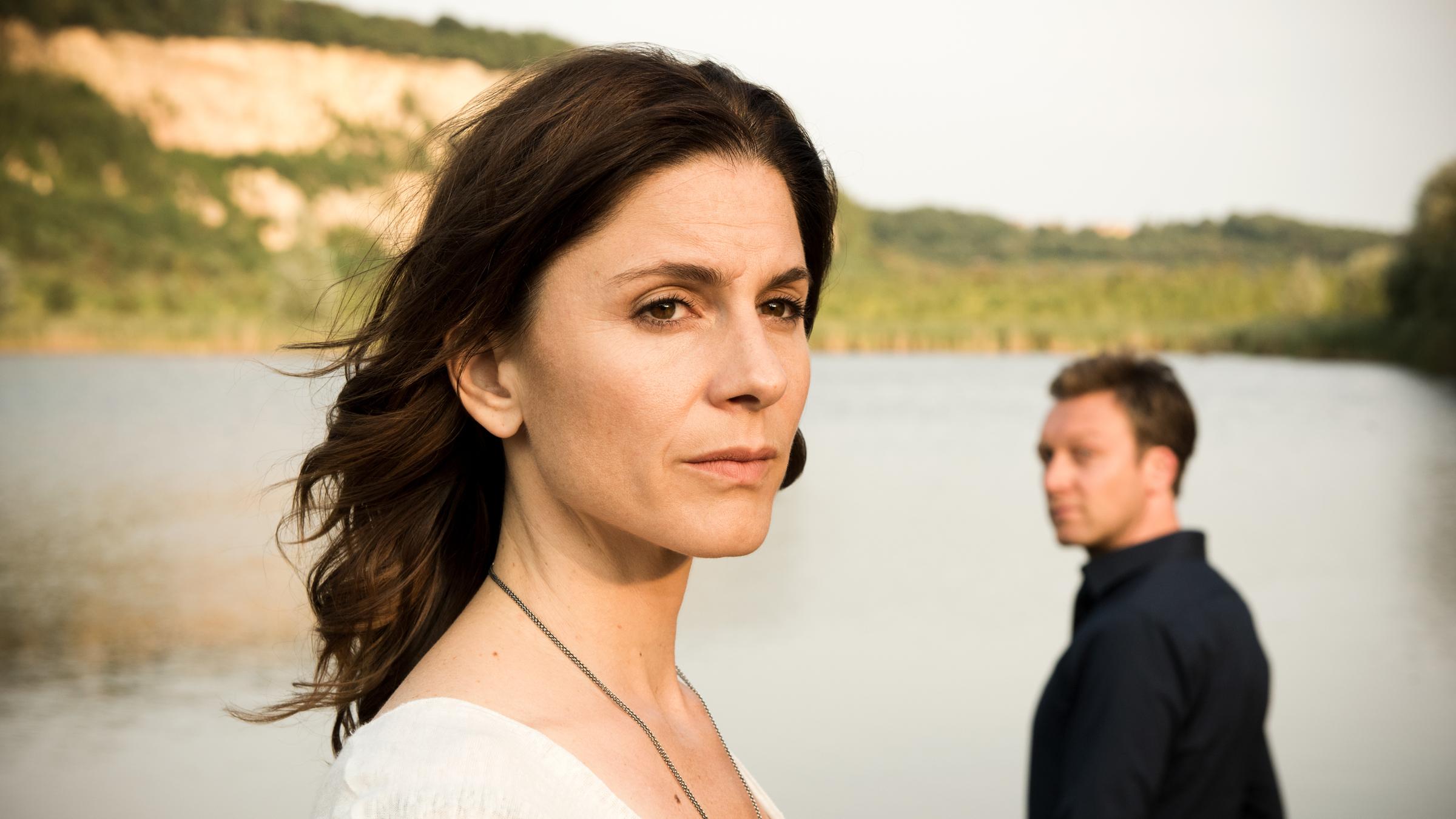 "In Wahrheit - Still ruht der See": Judith Mohn (Christina Hecke) blickt nachdenklich aus dem Bild. Hinter ihr steht Freddy Breyer (Robin Sondermann) und schaut zu Judith hin. Im Hintergrund ist ein See zu sehen.