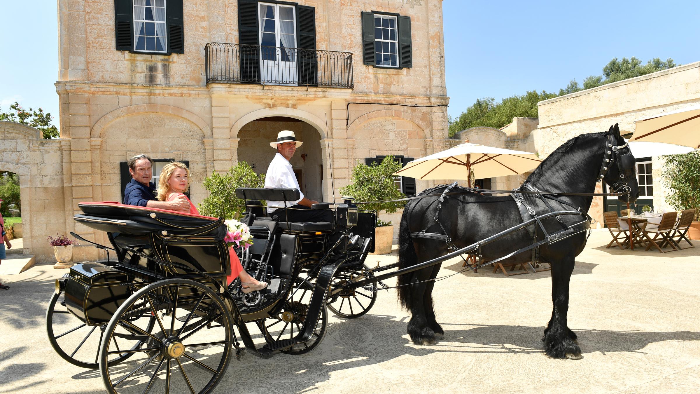 "Kreuzfahrt ins Glück - Hochzeitsreise nach Menorca": Iga (Karolina Lodyga) und Hans (Helmut Zierl) sitzen in einer schwarzen Kutsche, die vor einer Finca steht. Iga hält einen Blumenstrauß in den Händen.