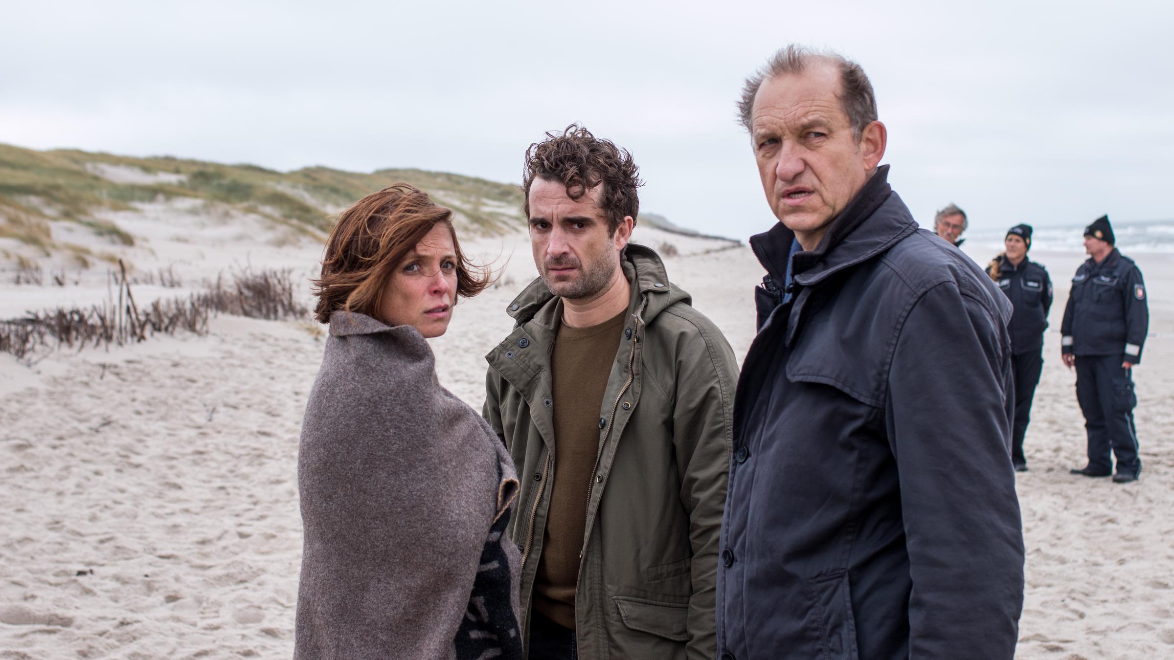 „Nord Nord Mord – Sievers und die tödliche Liebe“: Ina Behrendsen (Julia Brendler), Hinnerk Feldmann (Oliver Wnuk) und Carl Sievers (Peter Heinrich Brix) stehen am Strand und blicken in Richtung Kamera. Hinter ihnen stehen drei weitere Polizisten in Uniform.