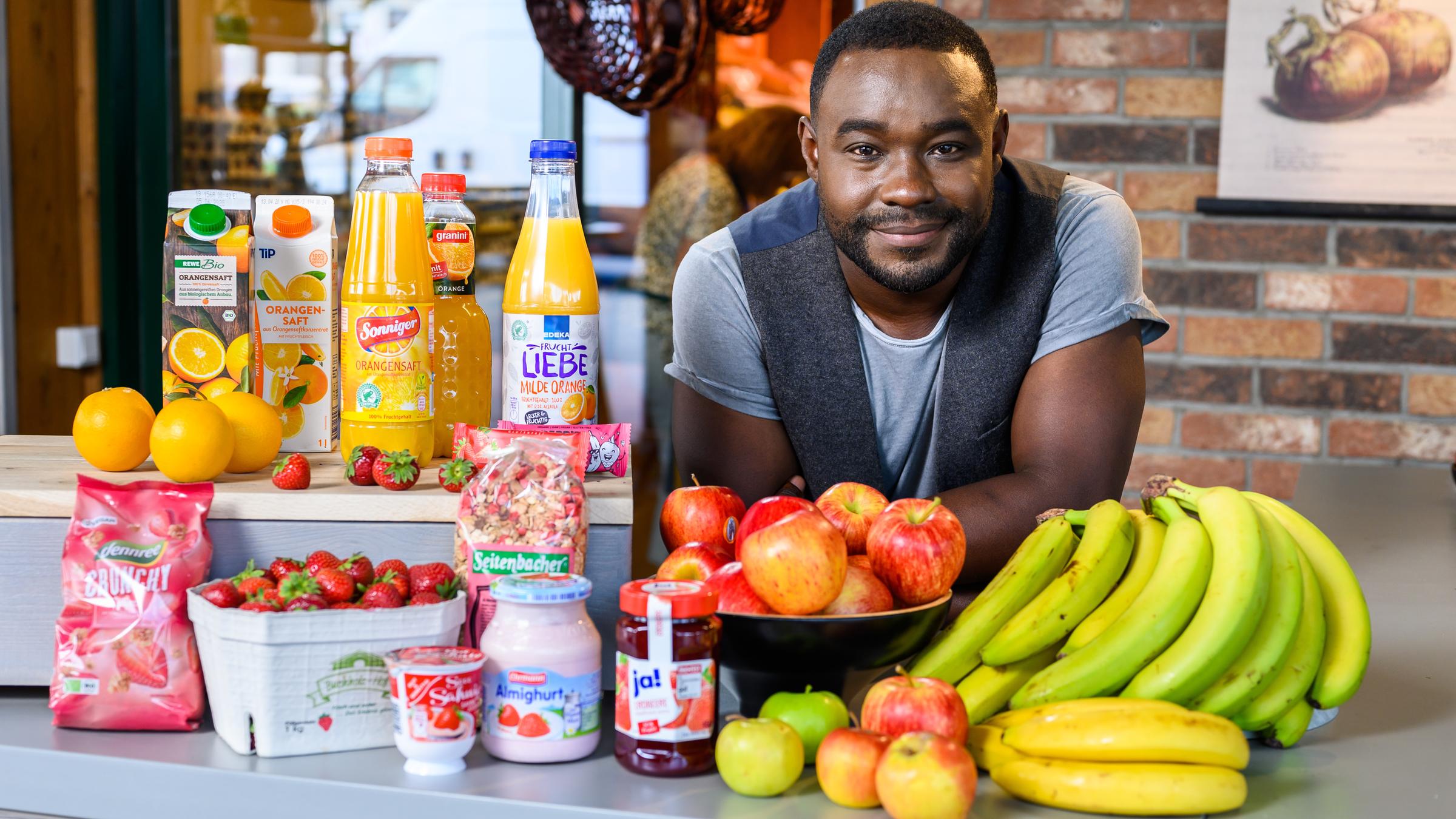 "ZDFzeit: Nelson Müllers Lebensmittelreport - Wie gut sind Äpfel, Orangensaft und Bio-Bananen?": Nelson Müller steht mit aufgelehnten Unterarmen an einem Tisch, auf dem Obst, Joghurt, Marmelade und Fruchtsäfte aufgebaut sind. Er blickt freundlich in Richtung Kamera.