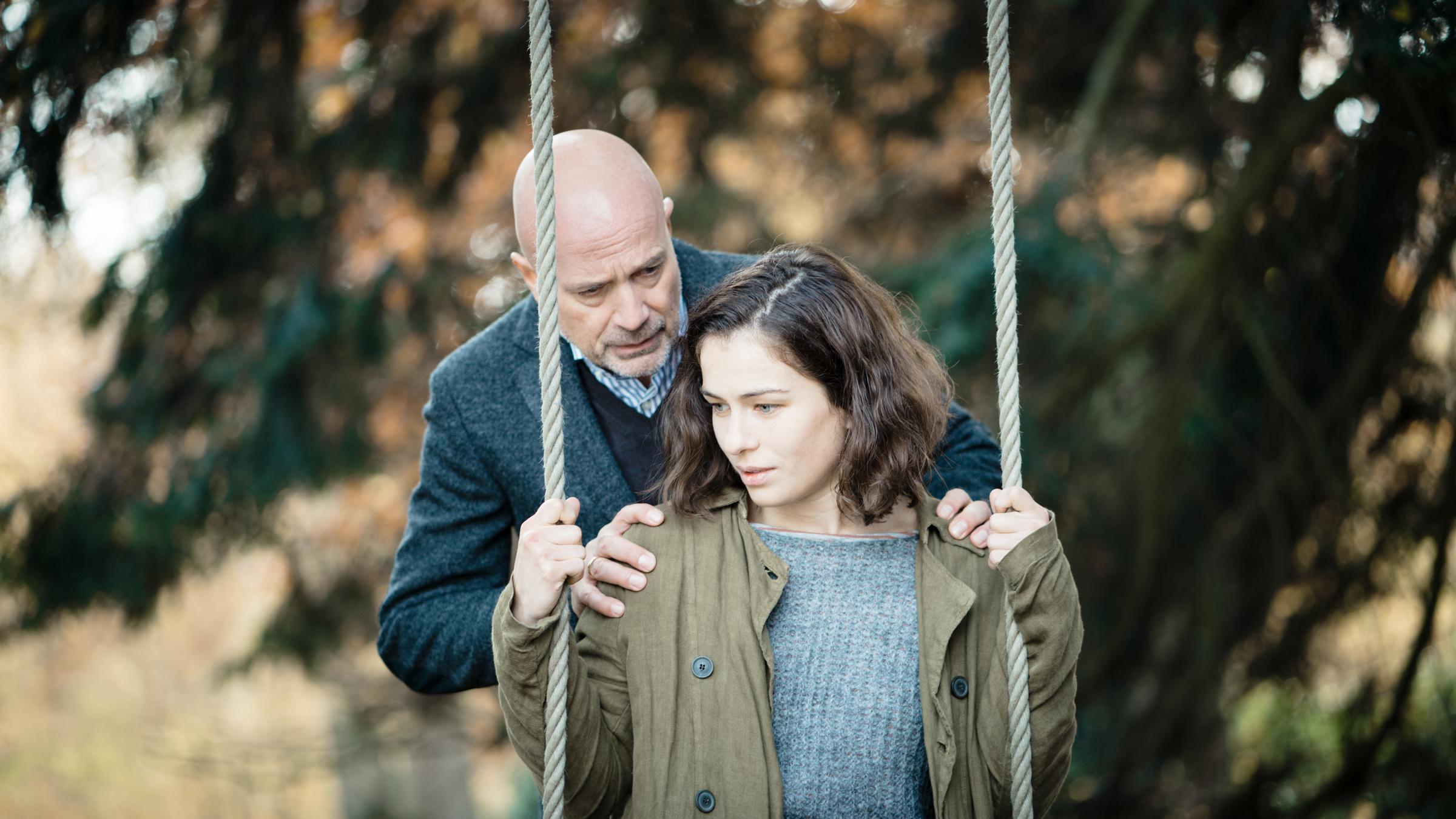"Die verlorene Tochter - Lügen": Isa von Gems (Henriette Confurius) sitzt auf der Schaukel im Garten. Hinter ihr steht ihr Vater Heinrich (Christian Berkel), der seine Hände auf ihre Schultern legt und mit ihr spricht.