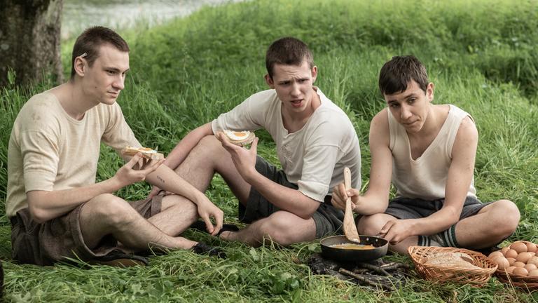 "Die Kinder von Windermere": Sam (Marek Wroblewski), Ike (Jakub Sprenger) und Arek (Tomasz Studzinski) sitzen im Wald auf einer Wiese und braten Eier.