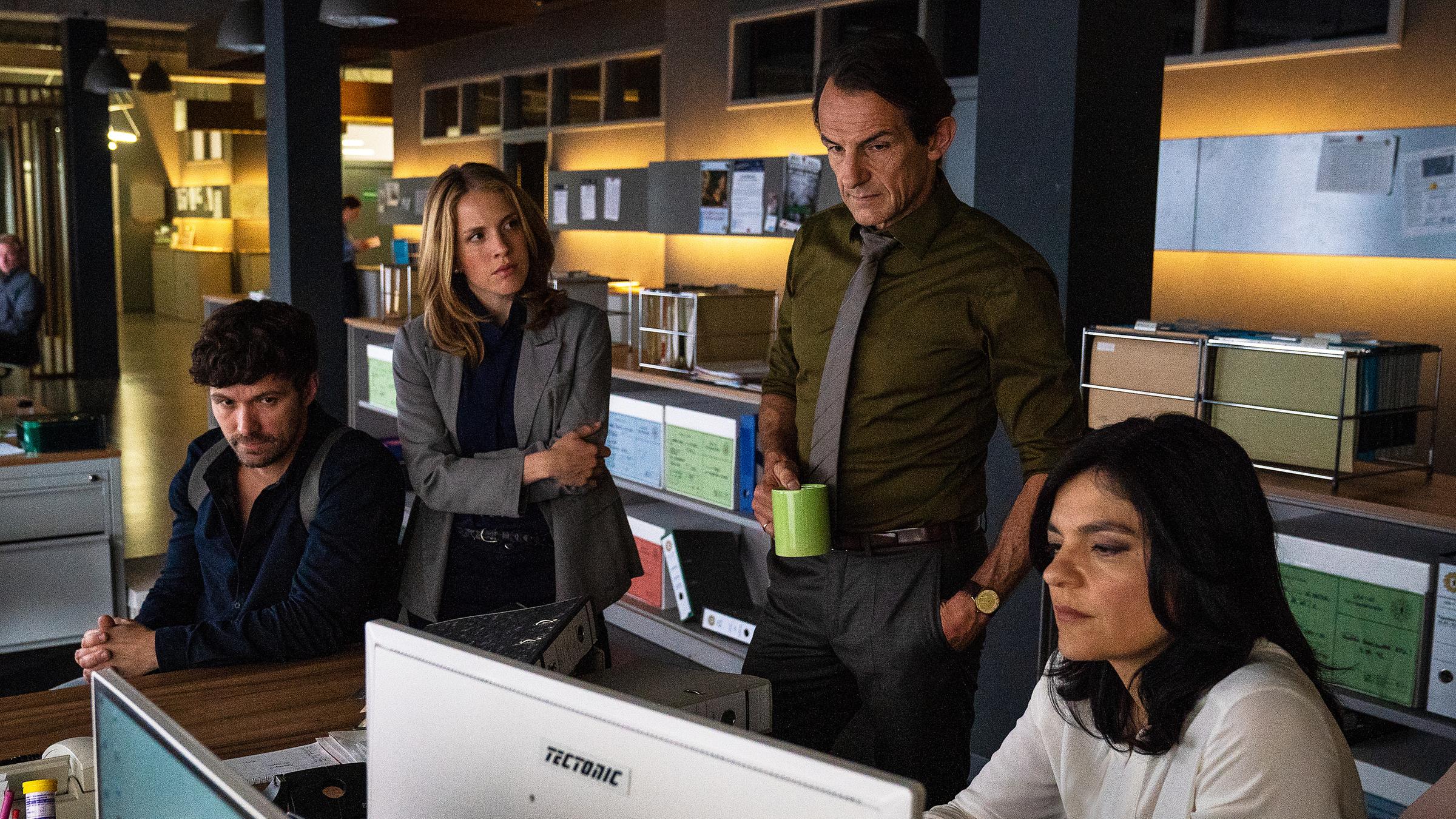 "Letzte Spur Berlin - Alibi": Mina Amiri (Jasmin Tabatabai) sitzt an ihrem Schreibtisch und schaut in den Bildschirm ihres Computers. Neben ihr steht Oliver Radek (Hans-Werner Meyer). Er hält eine Kaffeetasse in seiner rechten Hand und blickt ebenfalls auf den Monitor. Neben Radek steht Jessica Papst (Paula Kalenberg). Sie hat die Arme vor dem Oberkörper verschränkt und schaut zu Radek. Alex von Tal (Aleksander Radenkovic) sitzt am Kopfende des Schreibtisches auf einem Stuhl und blickt in Minas Richtung.