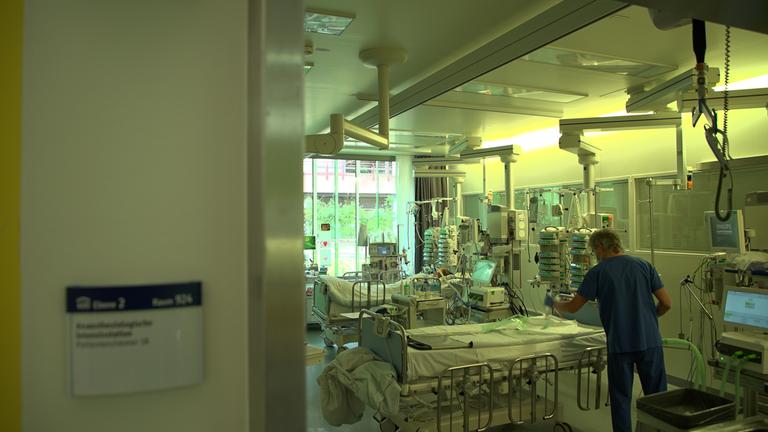 "ZDFzoom: Ärzte am Limit - Kostendruck statt Patientenwohl": Blick in ein Krankenzimmer mit zwei Krankenbetten. Am vorderen Bett steht ein Arzt in blauer Kleidung.