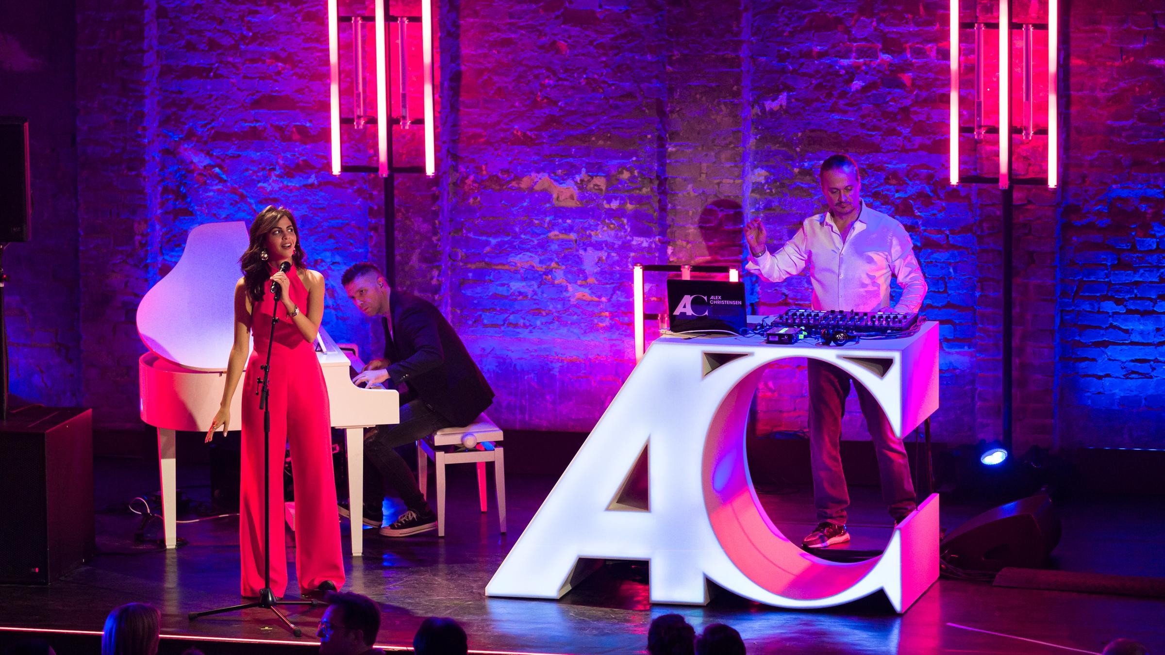 "Klassik im Club": Fatma Said steht auf der Bühne vor einem Mikrofon. Hinter ihr sitzt an einem weißen Klavier ein Pianist. Rechts steht Produzent Alex Christensen vor seinem DJ-Pult. Fatma Said schaut zu Alex Christensen.