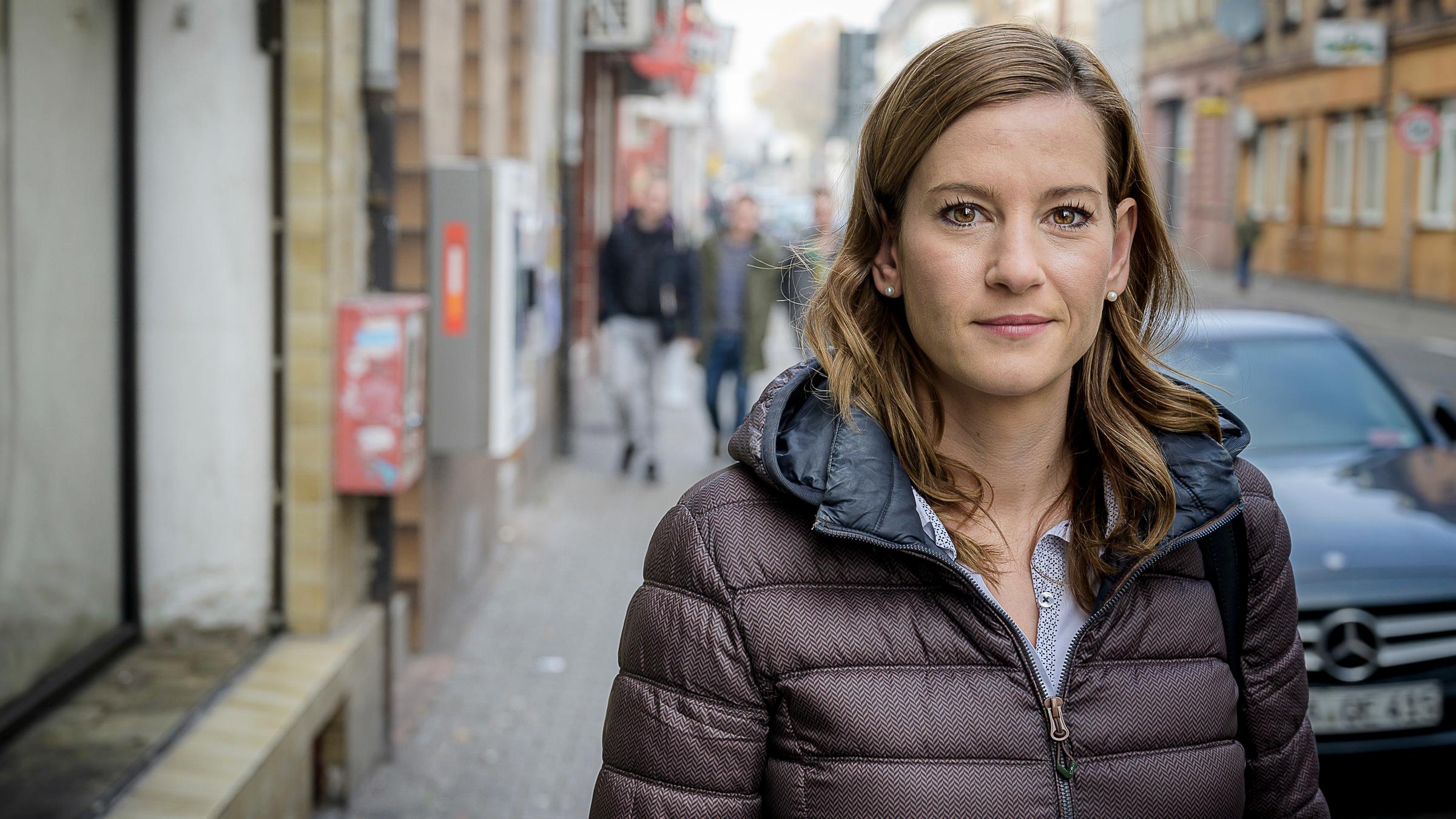 "37°: Zwei Quadratkilometer Stress - Hilfe für einen Stadtteil": Julia Wege, Gründerin des Projekts "Amalie" in der Mannheimer Neckarstadt, steht auf einem Bürgersteig und schaut in die Kamera. Im Hintergrund sieht man unscharf die Konturen von drei jungen Männern.