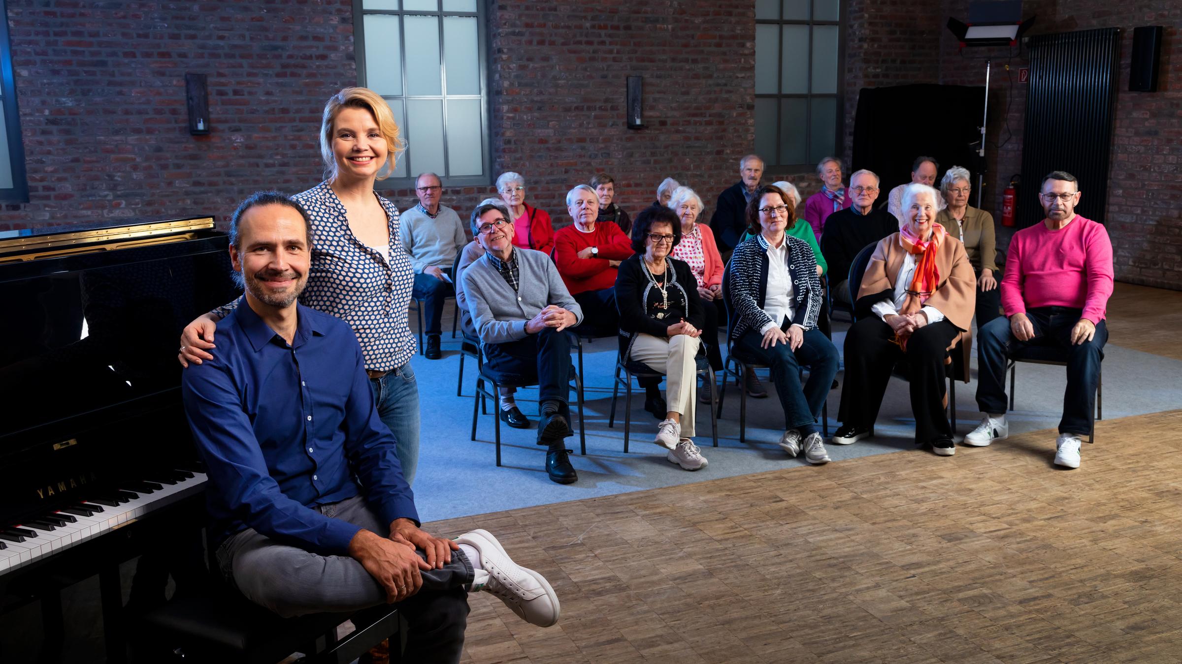 "Unvergesslich - Unser Chor für Menschen mit Demenz": Annette Frier und der Chorleiter Eddi Hüneke stehen vor einem Klavier und lachen in die Kamera. Im Hintergrund sind die Teilnehmer der Projektes zu sehen.