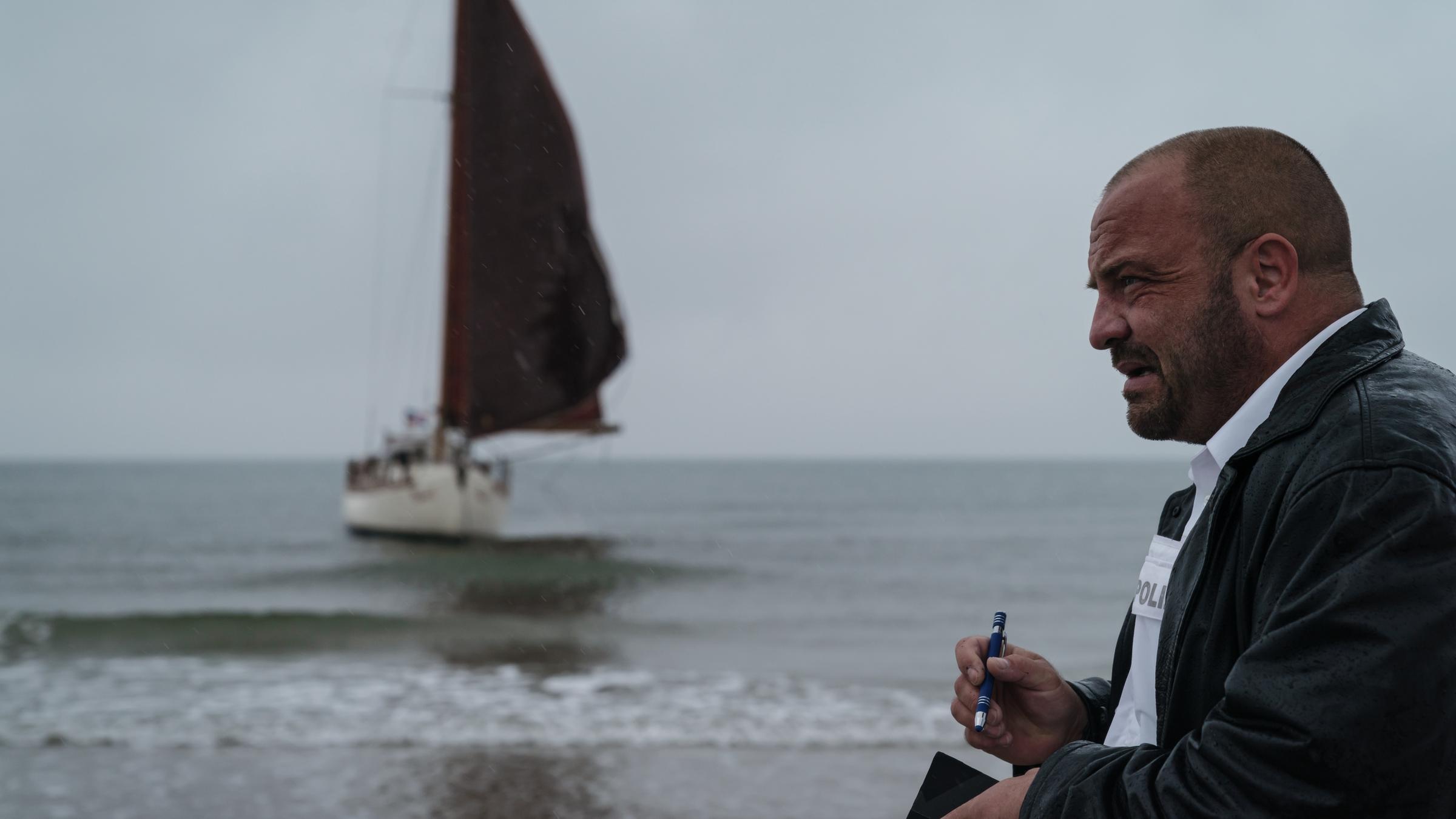"Sløborn": Polizist Mikkel Schwarting (Urs Rechn) steht mit Notizblock und Stift am Strand. Im Hintergrund sieht man die gestrandete Jacht.