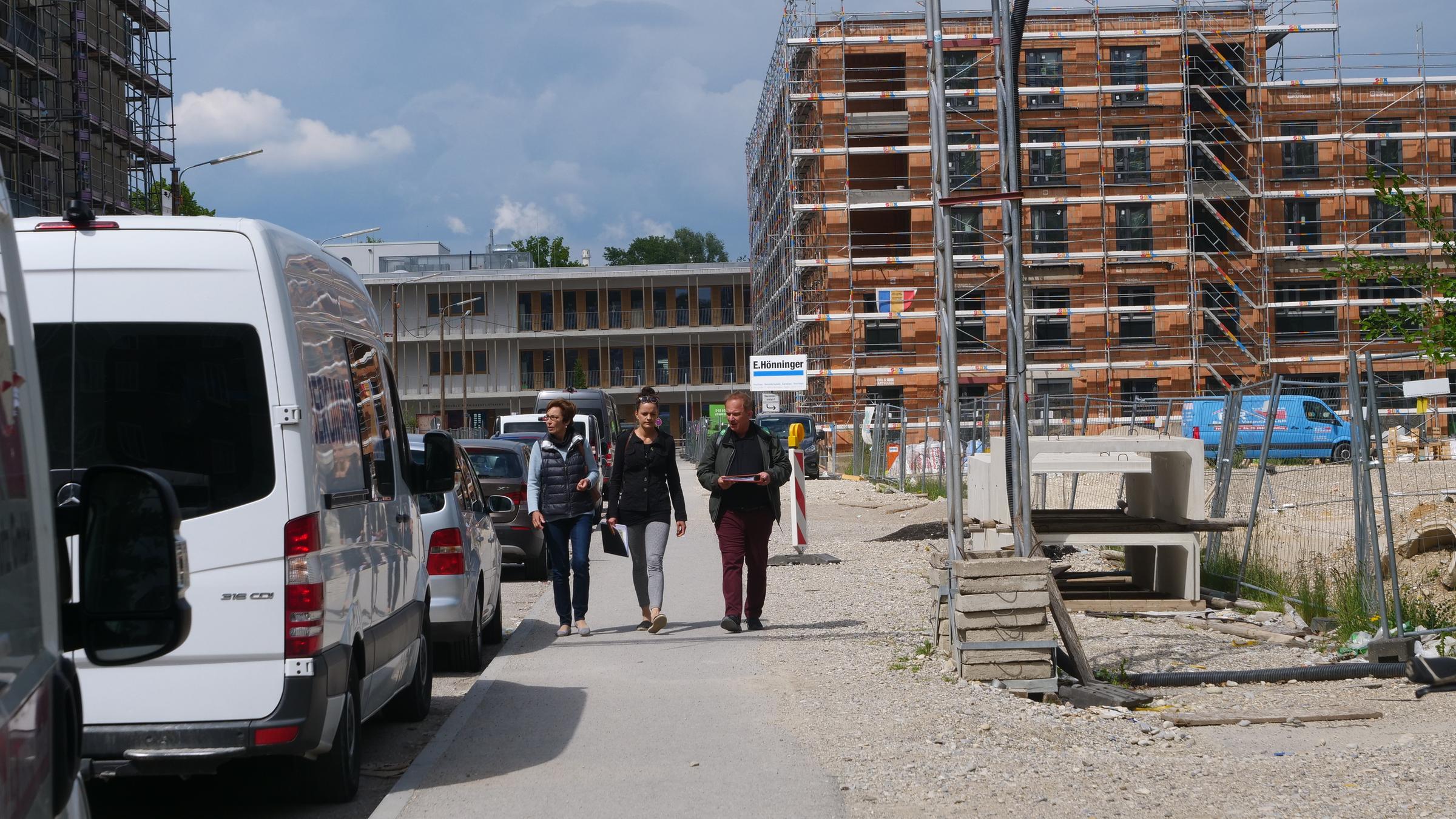 "plan b: Billiger Wohnen - Bessere Bedingungen für Mieter": Regina Lischka, Mara Roth und Claus Fincke gehen durch eine Neubausiedlung. Im Hintergrund ein mehrstöckiger Bau im Gerüst, am Bildrand links stehen Autos.