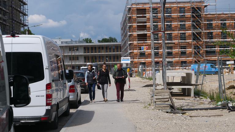 "plan b: Billiger Wohnen - Bessere Bedingungen für Mieter": Regina Lischka, Mara Roth und Claus Fincke gehen durch eine Neubausiedlung. Im Hintergrund ein mehrstöckiger Bau im Gerüst, am Bildrand links stehen Autos.