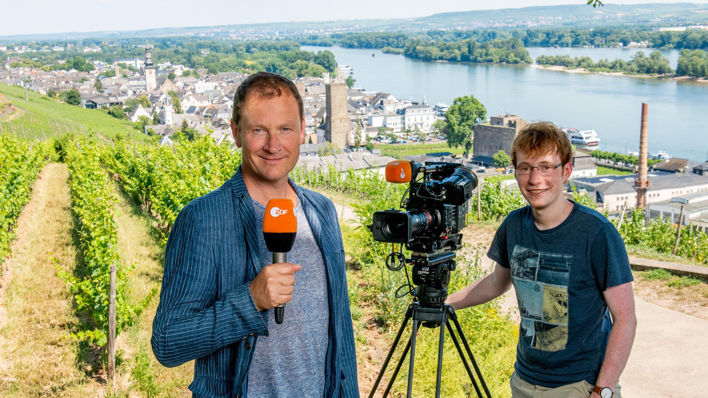 ZDF Reporter Peter Theisen mit Kameramann Christoph Timm stehen vor ländlicher Kulisse und lächeln in die Kamera.
