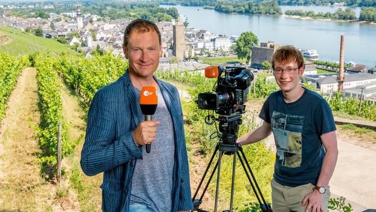 ZDF Reporter Peter Theisen mit Kameramann Christoph Timm stehen vor ländlicher Kulisse und lächeln in die Kamera.