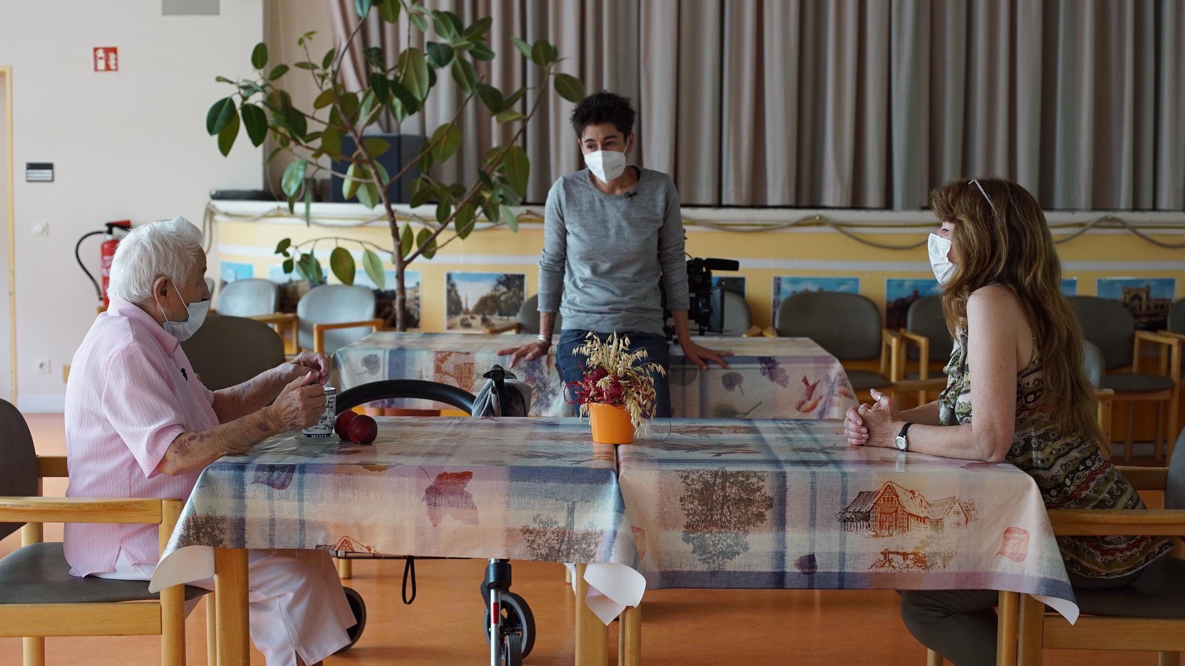 "dunja hayali", 16.7.2020: Dunja Hayali steht in einem Pflegheim an einem Tisch, an dem sich eine Pflegeheimbewohnerin und deren Enkeltochter gegenüber sitzen. Sie unterhalten sich. Alle drei tragen Mundschutzmasken.