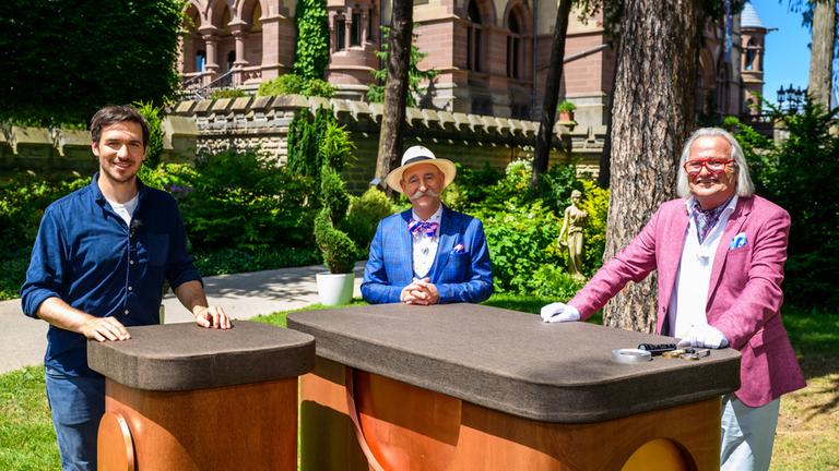 "Bares für Rares - Deutschlands größte Trödelshow": Felix Neureuther, Horst Lichter und Albert Maier stehen in großem Abstand zueinander hinter zwei Tischen und lächeln in die Kamera.