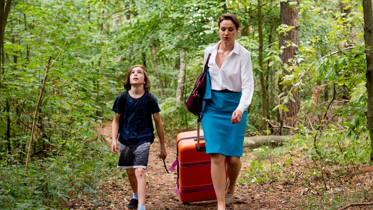 "Sterne über uns": Auf einem Waldweg zieht Melli (Franziska Hartmann) in einer Flugbegleiter Uniform einen großen orangefarbenen Trolley hinter sich her. Neben ihr läuft ihr neunjähriger Sohn Ben (Claudio Magno) und schaut staunend nach oben zu den Baumkronen.