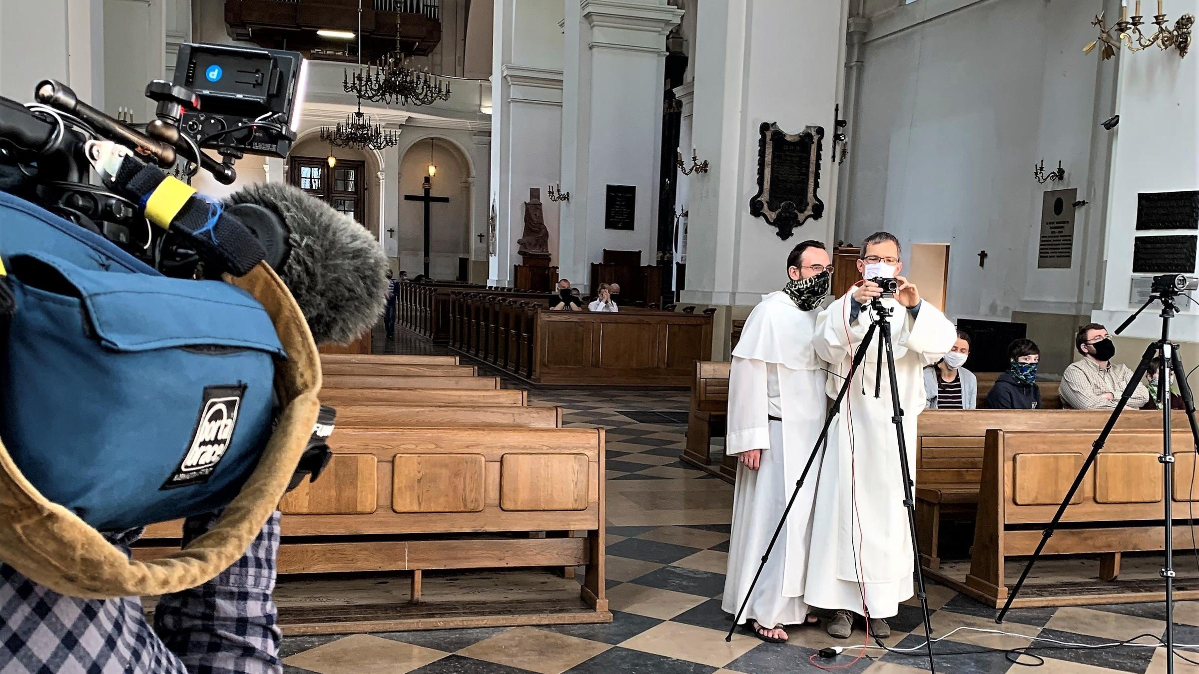 "Polen und die Pandemie - Kreativ in der Krise": Zwei Dominikanermönche stehen in einer Kirche zwischen fast leeren Bänken und hantieren mit Kameras, um den Gottesdienst online zu übertragen. Rechts im Bild ein Mann, der eine Kamera hält.