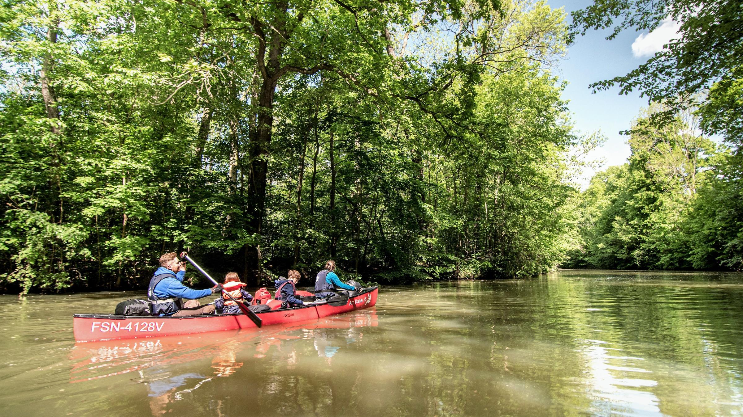 "Urlaub vor der Haustür": Eine vierköpfige Familie fährt in einem roten Kanu einen Fluss entlang, an dem hohe Bäume stehen.
