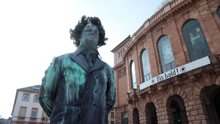 „Beethoven – Ein Geisterspiel“: Eine große Statue Ludwig van Beethovens steht vor dem Staatstheater Mainz. Die Statue ist dunkel gefärbt und hat einen weißen Fleck auf dem Gesicht. Die Augen der Statue sind geschlossen. Im Hintergrund ist das Staatstheater Mainz zu sehen, auf einem dort angebrachten Plakat steht „Bis bald“.