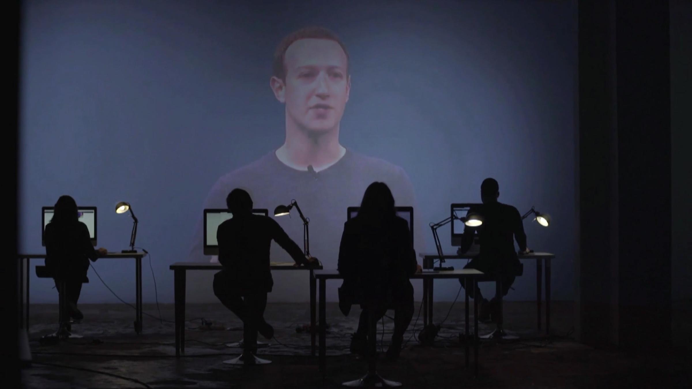 "Ghost Workers": Vier Menschen sitzen mit dem Rücken zum Betrachter an Computern. Auf einer großen Leinwand vor ihnen wird Mark Zuckerberg in Nahaufnahme gezeigt.