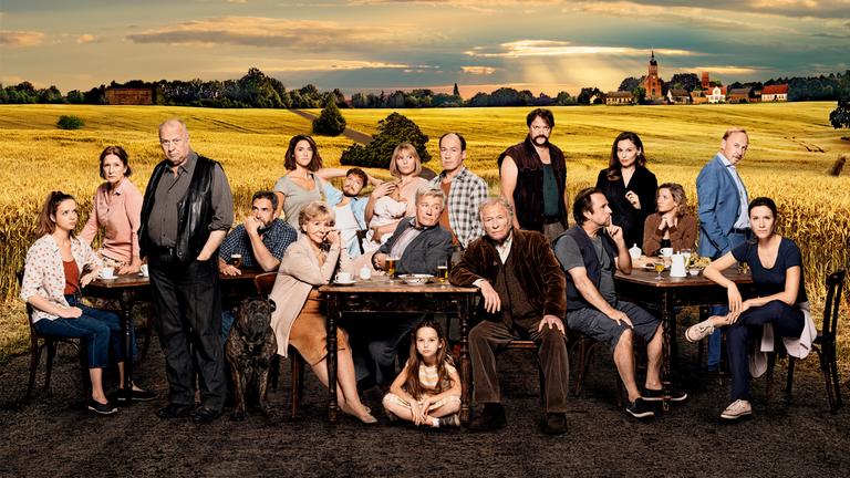 "Unterleuten - Das zerrissene Dorf" / Dreiteiler: Betty Kessler (Sarina Radomski), Hilde Kessler (Dagmar Manzel), Gombrowski (Thomas Thieme), Björn (Alexander Hörbe), Linda Franzen (Miriam Stein), Elena Gombrowski (Christine Schorn sitzend), Frederick (Jacob Matschenz), Jule Fließ (Rosalie Thomass), Arne Seidel (Jörg Schüttauf), Gerhard Fließ (Ulrich Noethen), Kron (Hermann Beyer), Schaller (Charly Hübner), Wolfi Hübschke (Bjarne Mädel), Anne Pilz (Mina Tander), Miriam Schaller (Nina Gummich), Meiler (Alexander Held), Kathrin Kron-Hübschke (Bettina Lamprecht), Krönchen (Una Lir, in der Bildmitte unter einem der Tische am Boden sitzend).