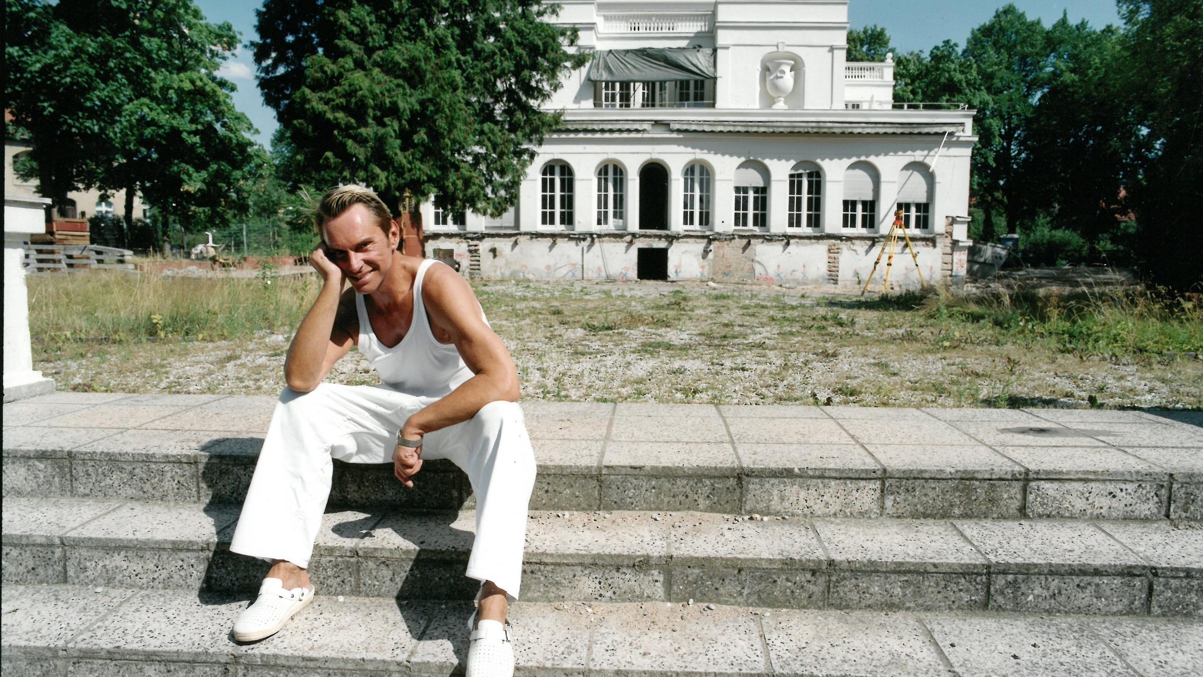 "Deutschlands große Clans - Die Joop-Story": Wolfgang Joop sitzt auf Treppenstufen, im Hintergrund die Villa Wunderkind in Potsdam.