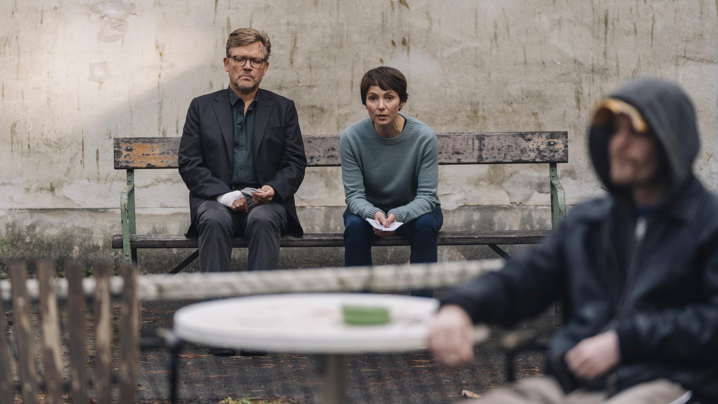 „Im Schatten der Angst“: Carsten Spanger (Justus von Dohnányi) und Karla Eckhardt (Julia Koschitz) sitzen draußen auf einer alten Bank. Carsten Spanger trägt einen Verband um seine rechte Hand, Karla Eckhardt hat einen Zettel in der Hand und im Vordergrund sieht man unscharf einen Mann mit einer Kapuze auf dem Kopf an einem Tisch sitzen.