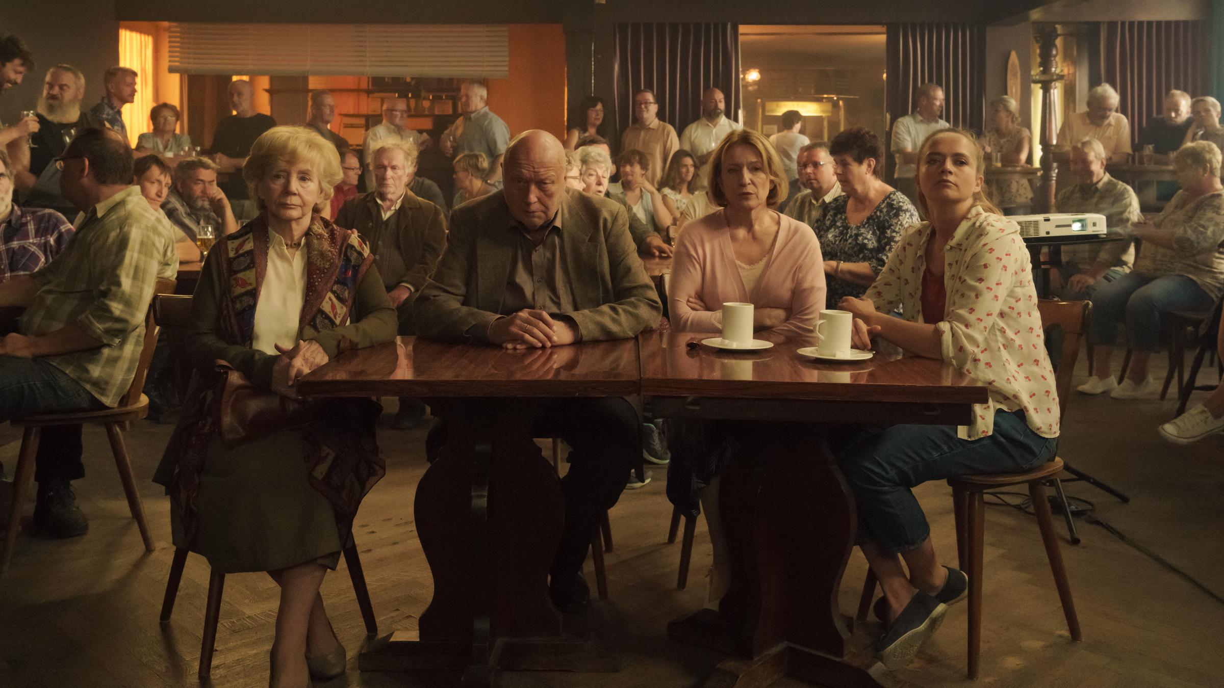 Unterleuten - Das zerrissene Dorf (1)": Rudolf Grombrowski (Thomas Thieme), Elena Grombrowski (Christine Schorn), Hilde Kessler (Dagmar Manzel) und Betty Kessler (Sarina Radomski) sitzen im Besprechungsraum vor allen anderen Leuten und schauen nach vorne auf die Projektvorstellung.