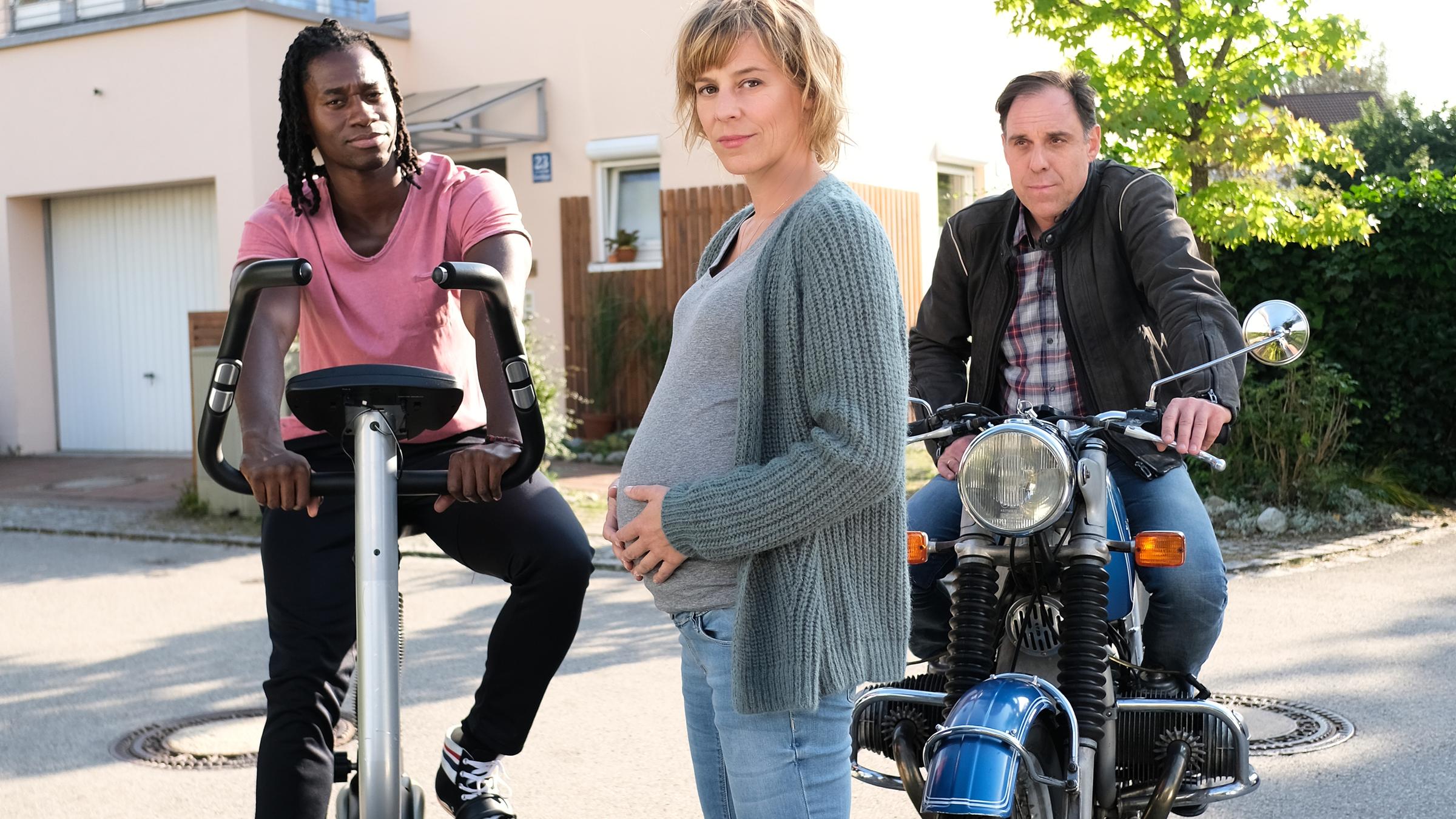  "Annie - kopfüber ins Leben": Annie (Bernadette Heerwagen) steht mit Babybauch vor ihrem Haus auf der Straße. Links neben ihr Hip-Hop-Trainer Raimund (Eugene Boateng), der auf einem Fitness-Bike sitzt. Rechts neben Annie sitzt ihr Mann Ralf (Thomas Loibl) auf einem alten Motorrad.