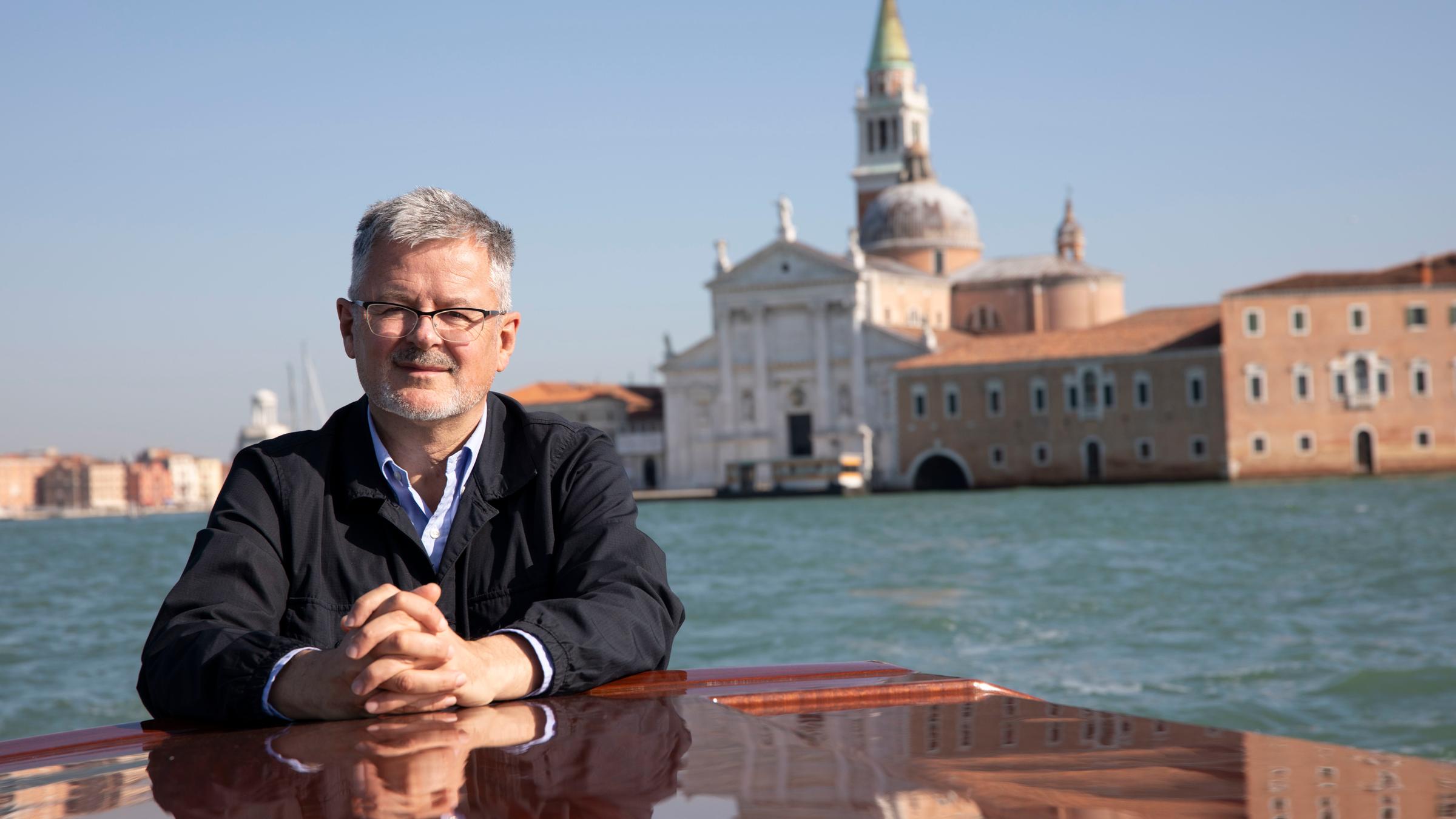 "Terra X: Welten-Saga - Die Schätze Europas": Christopher Clark ist auf einem Boot in der Lagune von Venedig. Er hat die Hände geschlossen auf das hölzerne Boot gelegt. Im Hintergrund ist der Markttum, der Kampanile, zu sehen.