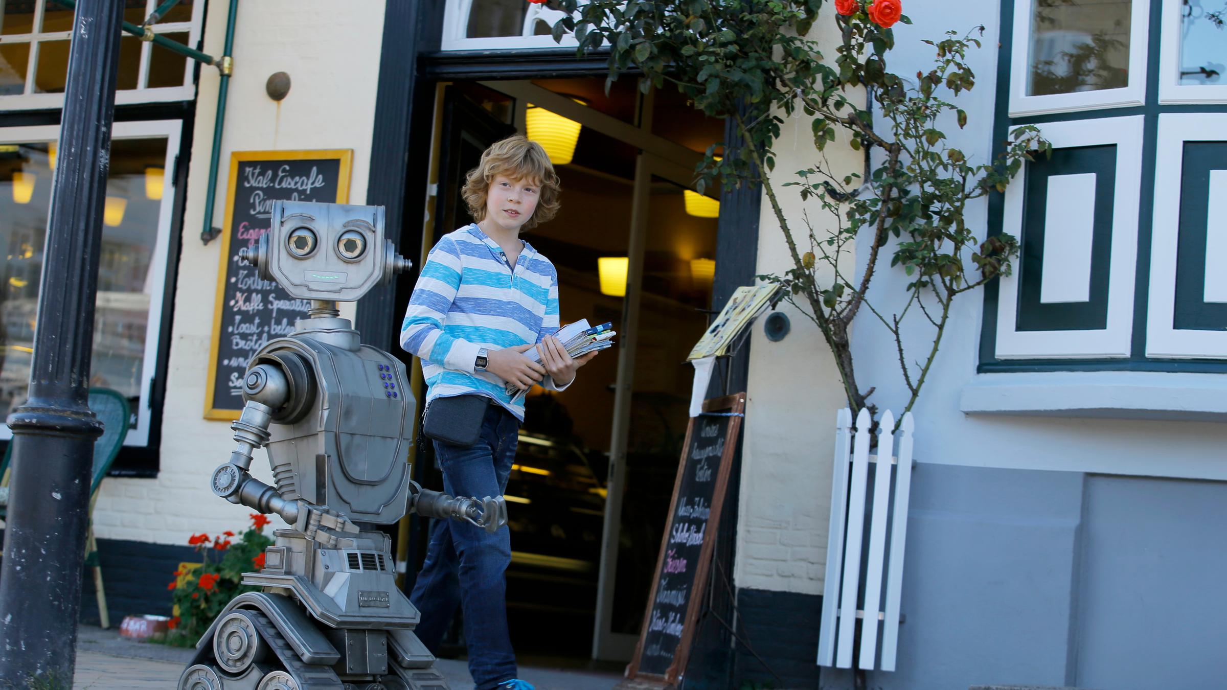 "Robbi, Tobbi und das Fliewatüüt": Tobbi (Arsseni Bultmann) und Roboter Robbi laufen beziehungsweise rollen nebeneinander die Straße entlang.